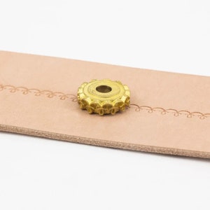 May include: A gold-colored metal stamp with a decorative design, featuring a circular shape with a central hole. The stamp is resting on a light brown leather surface.