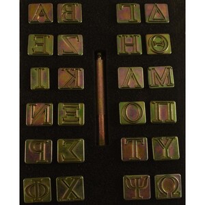 Puede incluir: Un juego de 24 sellos de letras de metal dorado en una caja negra forrada de espuma. Los sellos est&aacute;n dispuestos en dos filas de 12, con una herramienta de metal en el centro. Los sellos presentan el alfabeto griego, incluyendo letras como Alfa, Beta, Gamma, Delta, Epsilon, Zeta, Eta, Theta, Iota, Kappa, Lambda, Mu, Nu, Xi, Omicron, Pi, Rho, Sigma, Tau, Upsilon, Phi, Chi, Psi y Omega.