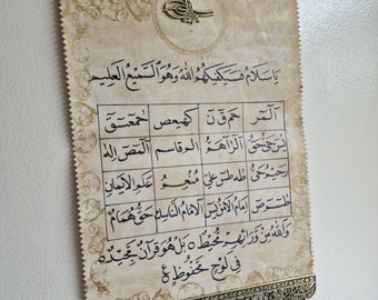 Può includere: Un arazzo decorativo da parete con una cornice in metallo decorata e nappe. L'arazzo presenta calligrafia araba in inchiostro nero su uno sfondo color crema. Il testo recita "Ya Salam, Fasiyakhum Allahu Wa Huwa As Sami'u Al 'Alim".