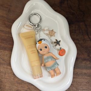 May include: A white, organically shaped ceramic dish holds a Laneige lip balm, a small doll, and a keychain with charms. The keychain features a penguin, a starfish, a ship's wheel, and an orange charm. The lip balm is vanilla-scented.
