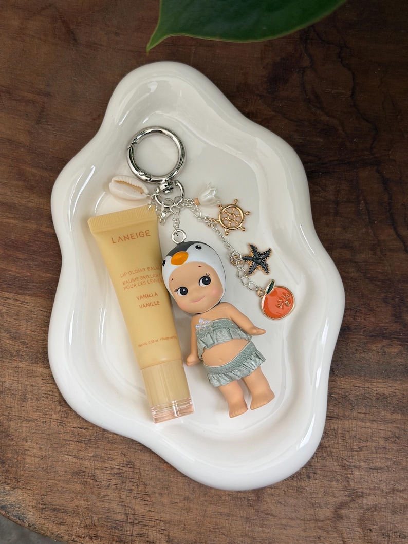 May include: A white, organically shaped ceramic dish holds a Laneige lip balm, a small doll, and a keychain with charms. The keychain features a penguin, a starfish, a ship's wheel, and an orange charm. The lip balm is vanilla-scented.