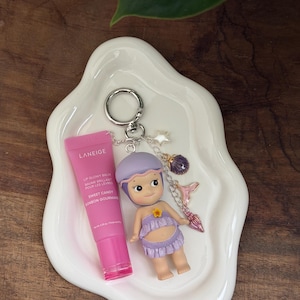 May include: A pink lip gloss tube and a keychain with a doll figure and charms rest on a white, wavy-edged dish. The doll wears a purple outfit. The keychain includes a star, a purple gem, and a pink mermaid tail charm.