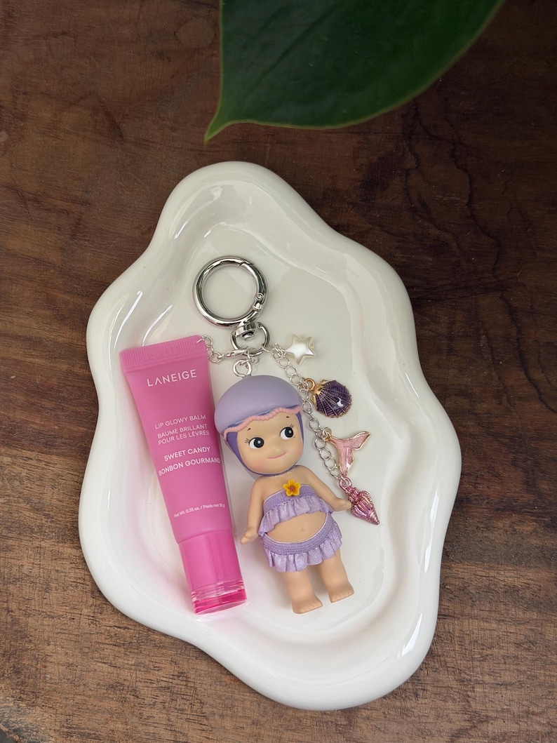 May include: A pink lip gloss tube and a keychain with a doll figure and charms rest on a white, wavy-edged dish. The doll wears a purple outfit. The keychain includes a star, a purple gem, and a pink mermaid tail charm.
