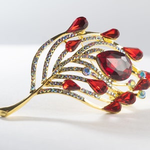 May include: A gold-toned brooch in a leaf design, adorned with red teardrop-shaped and oval-cut gemstones. The brooch also features iridescent, clear crystals, creating a sparkling effect. The jewelry piece is set against a white background.