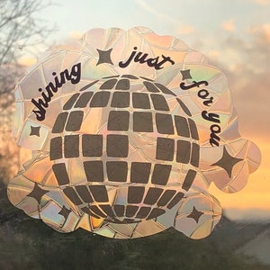 May include: A holographic sticker with a disco ball design and the words "shining just for you". The sticker has a scalloped edge and star accents. The background shows a sunset.