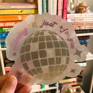 May include: A clear sticker with a disco ball design. The sticker has a scalloped edge and features the words "shining just for you" in pink script. Gold star accents surround the disco ball.