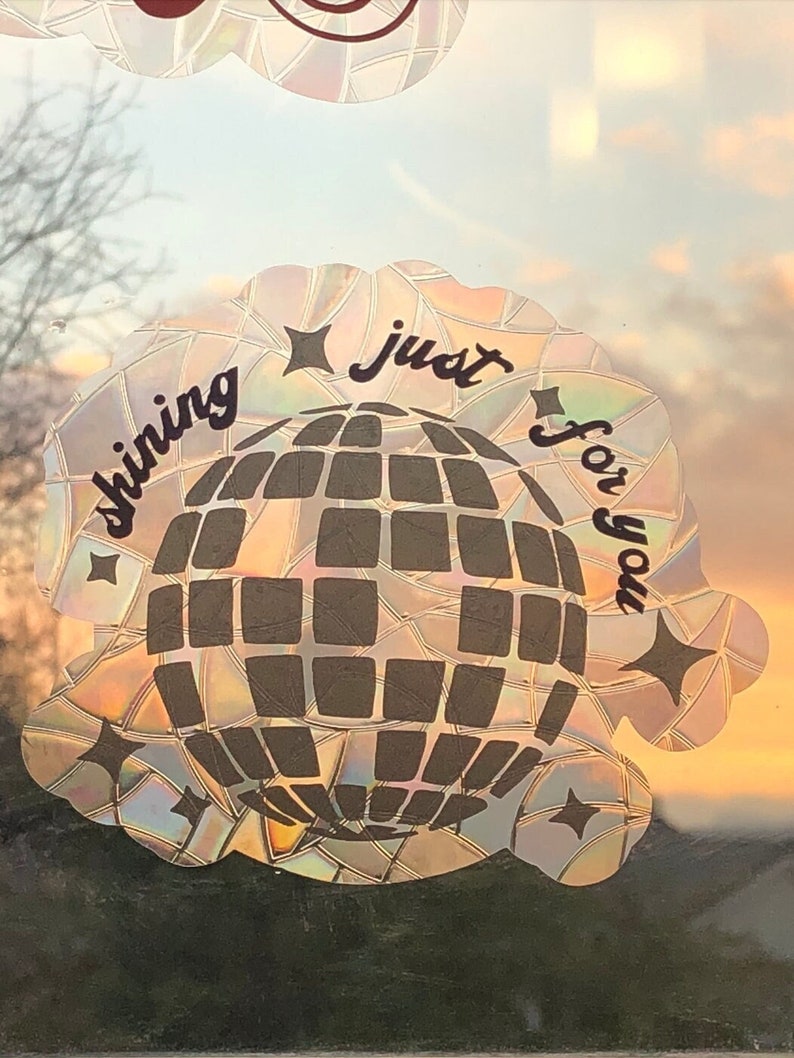 May include: A holographic sticker with a disco ball design and the words "shining just for you". The sticker has a scalloped edge and star accents. The background shows a sunset.