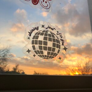 May include: Window stickers featuring a disco ball and smiley faces. The disco ball sticker has the text "shining just for you". The smiley face stickers are red on a clear background. The stickers are against a sunset background.