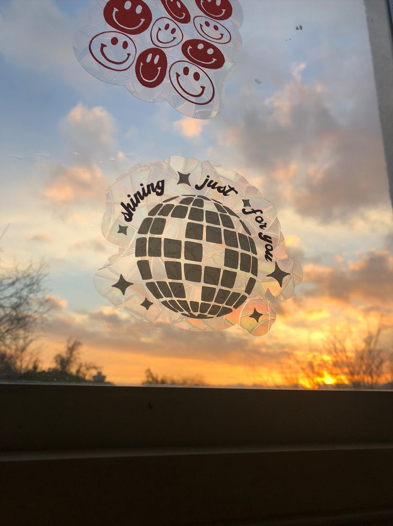 May include: Window stickers featuring a disco ball and smiley faces. The disco ball sticker has the text "shining just for you". The smiley face stickers are red on a clear background. The stickers are against a sunset background.