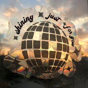 May include: A holographic sticker with a disco ball design and the text "shining just for you." The sticker has a reflective, iridescent finish, with a black disco ball pattern and star accents. The background shows a sunset.