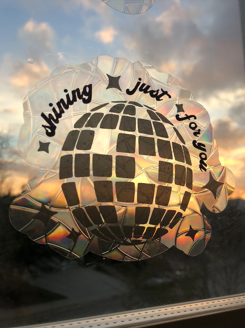 May include: A holographic sticker with a disco ball design and the text "shining just for you." The sticker has a reflective, iridescent finish, with a black disco ball pattern and star accents. The background shows a sunset.