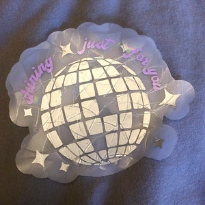 May include: A holographic sticker with a disco ball design. The sticker has the words "shining just for you" in a light purple script. The disco ball is white and surrounded by star shapes.