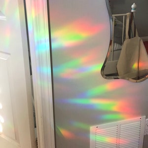 May include: A wall with a white door frame and a white vent, illuminated by a rainbow light effect. The light creates streaks of red, orange, yellow, green, and blue across the wall. A mirror with a unique shape is visible.
