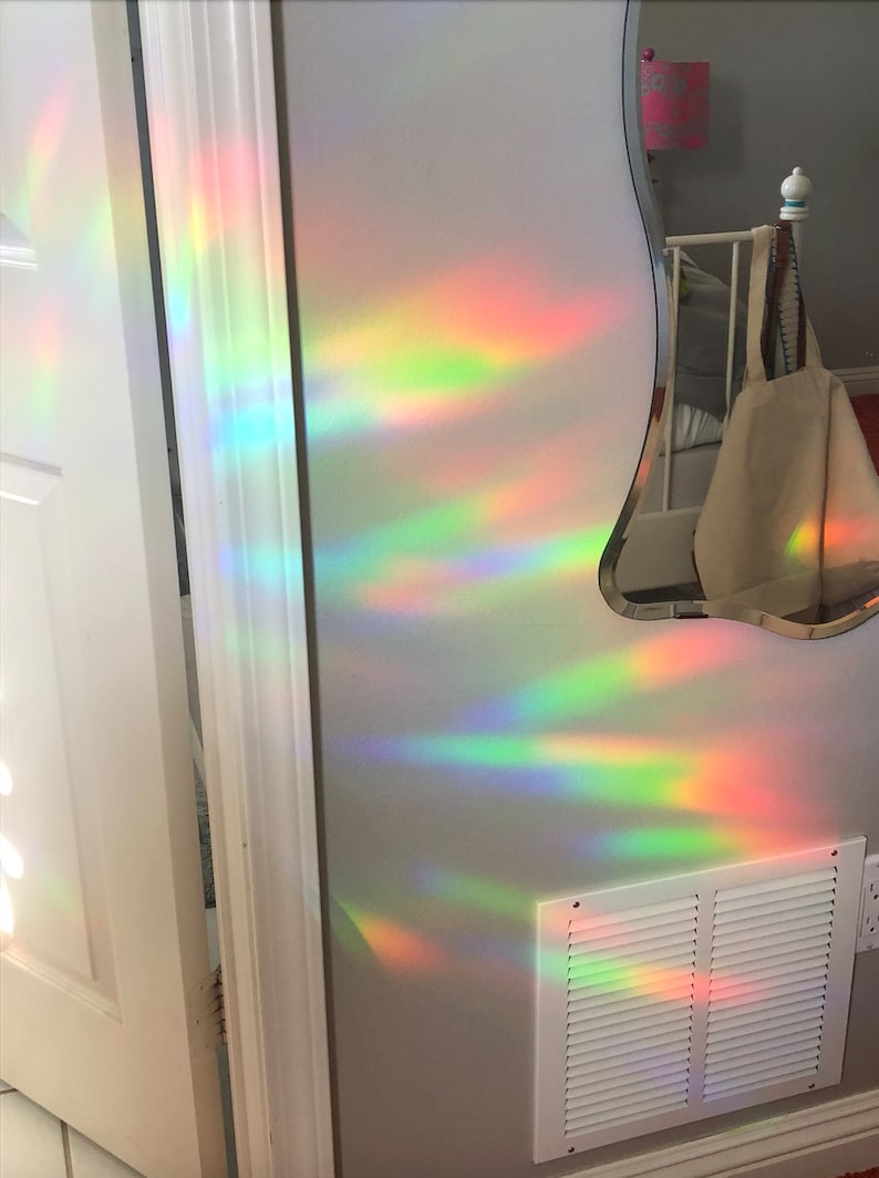 May include: A wall with a white door frame and a white vent, illuminated by a rainbow light effect. The light creates streaks of red, orange, yellow, green, and blue across the wall. A mirror with a unique shape is visible.