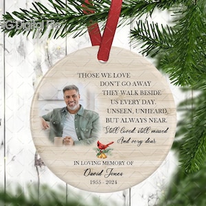 Pode incluir: Um ornamento redondo de madeira com fundo branco e texto preto. O texto diz "Those we love don't go away they walk beside us every day. Unseen, unheard, but always near. Still loved, still missed and very dear. In loving memory of David Jones 1955-2024". Um cardeal vermelho com um ramo de azevinho vermelho e verde está no canto inferior esquerdo. Uma foto de um homem está no canto superior direito.
