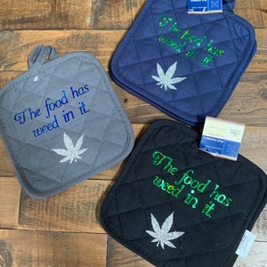 May include: Three quilted pot holders in gray, navy blue, and black. Each pot holder features the text "The food has weed in it" and a glittery cannabis leaf design. The pot holders have a loop for hanging.