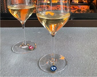 May include: Two stemmed wine glasses filled with white wine, each with a charm on the base. The background features a fireplace with a warm, glowing fire. The glasses are clear, and the wine has a golden hue.