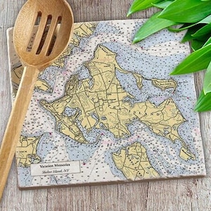 May include: A nautical map of Shelter Island, NY, printed on a ceramic tile. The tile is beige with a light blue and yellow map design. The text "Vacation Memories" and "Shelter Island, NY" is printed on the tile.