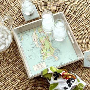 May include: A white wooden tray with a nautical map design featuring the text "Casco Bay" and "New England Summer" with a floral patterned fabric napkin on top.
