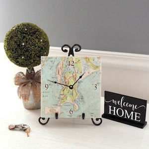 May include: A decorative clock with a map design on a black metal stand. The clock face has black hands and Roman numeral markings. The clock is sitting on a white surface with a small green topiary and a black sign that says "welcome home".