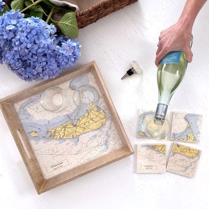 May include: A wooden serving tray and coasters feature nautical chart designs. A bottle of wine is being poured into a glass. Blue hydrangeas and two clear glasses are on the tray. The coasters and tray have the names "Charlene & James" and the date "August 6, 2022".