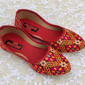 May include: A pair of red flat shoes with a colorful floral pattern. The shoes have a pointed toe and are embroidered with a variety of colors, including orange, pink, green, and white. The shoes have the text "ARTISANS TOUCH" printed on the inside.