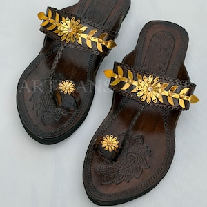 May include: A pair of dark brown leather sandals featuring embossed floral patterns on the footbed. The straps are adorned with metallic gold leaf-shaped accents and a central flower design. A small matching flower sits on the toe loop.