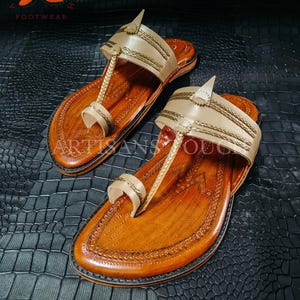 May include: A pair of handcrafted leather sandals with a tan upper and a brown sole. The sandals feature intricate stitching and a toe loop design. The sandals are displayed on a textured black surface. The logo "ARTISANS TOUCH FOOTWEAR" is in the top left corner.
