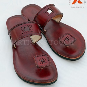 May include: Burgundy leather sandals with decorative cutouts and braided trim. The sandals have a toe loop and a strap across the top of the foot. The brand logo "ARTISANS TOUCH FOOTWEAR" is visible in the upper right corner.