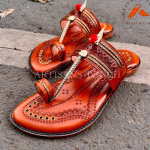 May include: A pair of handcrafted, brown leather sandals with intricate embossed designs. The sandals feature a toe loop, decorative straps, and a rope-like thong. Accented with gold trim and small red pom-poms. The text "ARTISANS TOUCH FOOTWEAR" is visible.