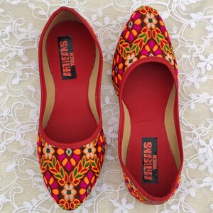 May include: A pair of red embroidered flat shoes with a floral pattern in shades of orange, pink, green, and white. The shoes have a pointed toe and a flat sole. The inside of the shoes has the text "ARTISANS TOUCH" printed on the insole.