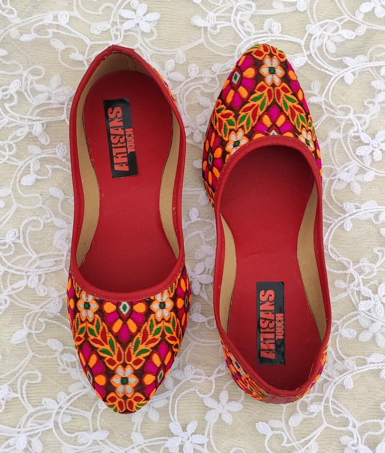 May include: A pair of red embroidered flat shoes with a floral pattern in shades of orange, pink, green, and white. The shoes have a pointed toe and a flat sole. The inside of the shoes has the text "ARTISANS TOUCH" printed on the insole.