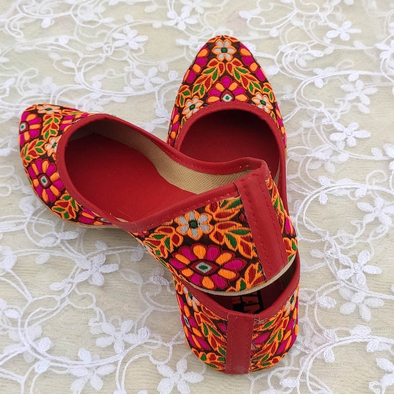 May include: A pair of red embroidered flat shoes with a floral pattern in shades of orange, pink, green, and white. The shoes have pointed toes and are made of a soft fabric.