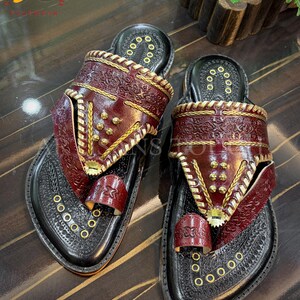 May include: Pair of handcrafted leather sandals. The sandals feature a black base with intricate embossed patterns and burgundy straps adorned with gold-colored studs and stitching. The toe strap and upper straps are burgundy.