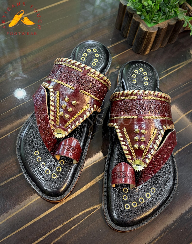 May include: Pair of handcrafted leather sandals. The sandals feature a black base with intricate embossed patterns and burgundy straps adorned with gold-colored studs and stitching. The toe strap and upper straps are burgundy.