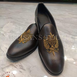 Op de afbeelding: Een paar bruine leren loafers met goud geborduurde wapenschilden. De schoenen hebben een ronde neus en een lage hak. Het logo "ARTISANS TOUCH FOOTWEAR" is zichtbaar onderaan de afbeelding.