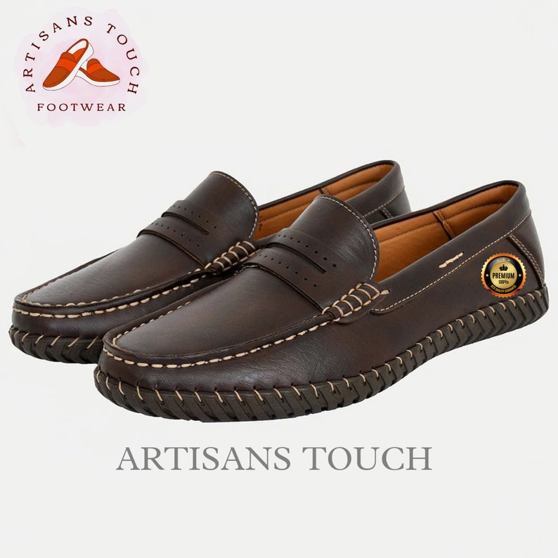 May include: A pair of brown leather loafers with a stitched design around the sole. The loafers have a  stitched detail across the top and a small, round, gold label with the text "PREMIUM 100%" on the right shoe. The text "ARTISANS TOUCH FOOTWEAR" is visible in a circle above the shoes.