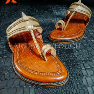 May include: A pair of handcrafted leather sandals with a light tan upper and a brown footbed. The sandals feature intricate stitching and a toe loop design. The words "ARTISANS TOUCH" are visible on the sandals. The sandals are displayed on a textured black surface.