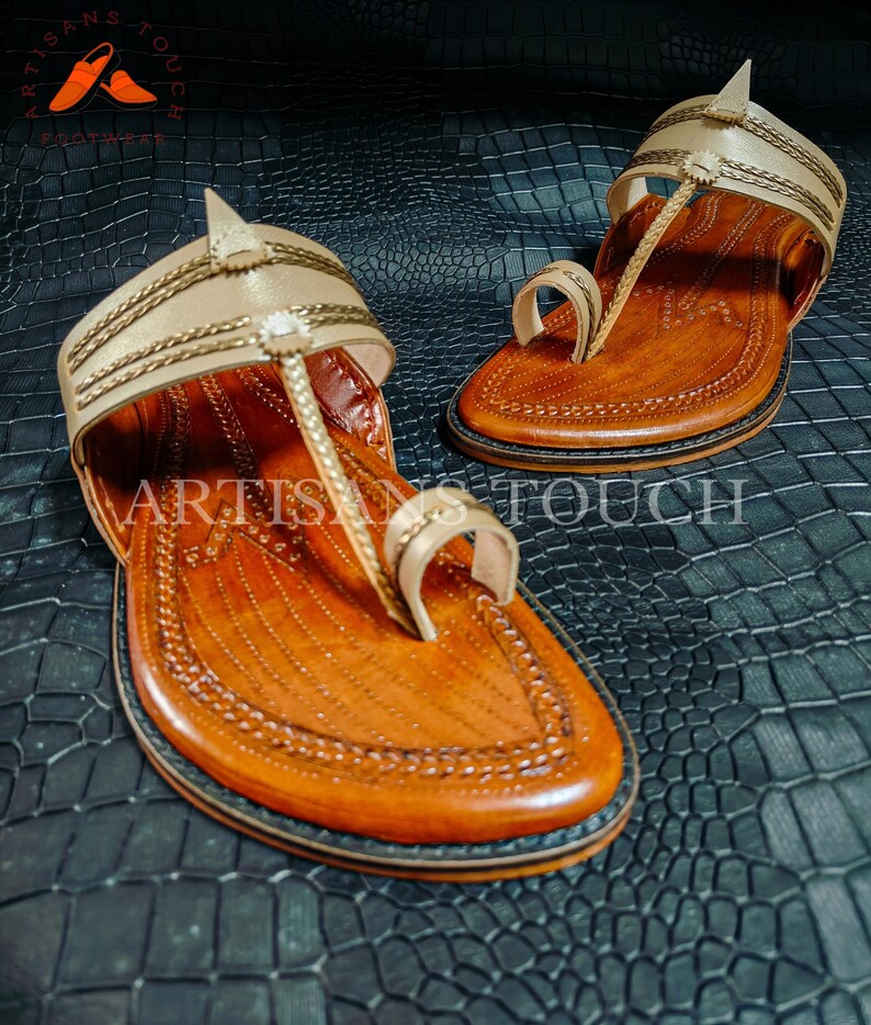 May include: A pair of handcrafted leather sandals with a light tan upper and a brown footbed. The sandals feature intricate stitching and a toe loop design. The words "ARTISANS TOUCH" are visible on the sandals. The sandals are displayed on a textured black surface.
