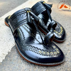 May include: Pair of black leather sandals with intricate embossed patterns and decorative gold-colored studs. The sandals feature braided straps, tassels, and a toe loop design. The sole has a brown edge. The sandals are labeled "ARTISANS TOUCH FOOTWEAR".