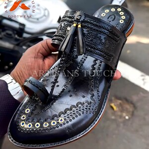 May include: Black leather sandal with intricate embossed floral and geometric patterns. Features a toe loop, decorative gold-colored eyelets, and tassel accents. The sandal is part of the Artisans Touch Footwear collection.