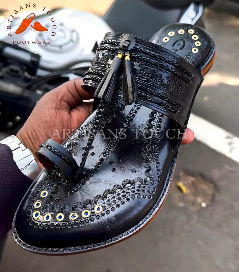 May include: Black leather sandal with intricate embossed floral and geometric patterns. Features a toe loop, decorative gold-colored eyelets, and tassel accents. The sandal is part of the Artisans Touch Footwear collection.