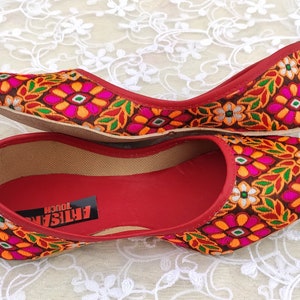 May include: A pair of red flat shoes with a floral embroidered design. The shoes have a brown sole and a red lining. The text "ARTISAN TOUCH" is visible on the inside of the shoe.