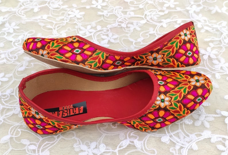 May include: A pair of red flat shoes with a floral embroidered design. The shoes have a brown sole and a red lining. The text "ARTISAN TOUCH" is visible on the inside of the shoe.