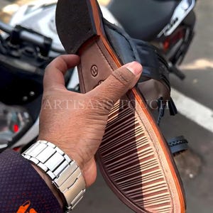 May include: Black and brown leather sandal with a woven strap detail. The sole is brown with a textured pattern. The sandal is held in a person's hand, with a silver watch visible. The text "ARTISANS TOUCH" is visible.