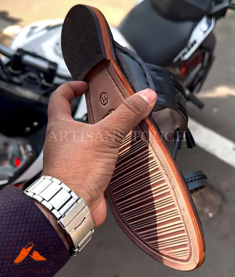 May include: Black and brown leather sandal with a woven strap detail. The sole is brown with a textured pattern. The sandal is held in a person's hand, with a silver watch visible. The text "ARTISANS TOUCH" is visible.