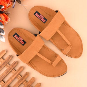 Op de afbeelding: Een paar bruine leren sandalen met een band over de bovenkant en een teenlus. De sandalen hebben de tekst "ARTISANS TOUCH" op de band.