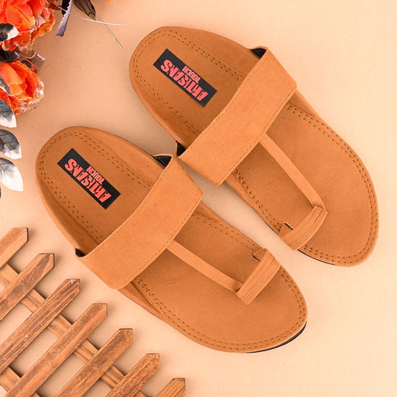 Op de afbeelding: Een paar bruine leren sandalen met een band over de bovenkant en een teenlus. De sandalen hebben de tekst "ARTISANS TOUCH" op de band.