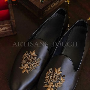 Op de afbeelding: Een paar zwarte leren loafers met gouden geborduurd bloemen- en wapenontwerp. De schoenen hebben een ronde neus en een instapmodel. De tekst "ARTISANS TOUCH" is zichtbaar.