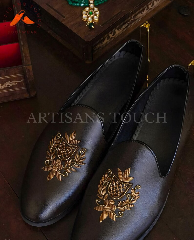 Op de afbeelding: Een paar zwarte leren loafers met gouden geborduurd bloemen- en wapenontwerp. De schoenen hebben een ronde neus en een instapmodel. De tekst "ARTISANS TOUCH" is zichtbaar.